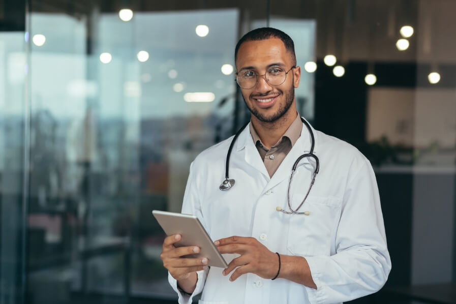 De fondo se ve distorsionado de un recibidor y en primer plano se ve un médico sonriendo con una tablet en la mano. 