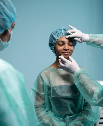 Se ve una paciente sentada vestida con ropa quirúrgica y se ven las manos cubiertas de látex de un médico tocando su rostro, además frente a ella hay otra médica mirando lo que está sucediendo. 