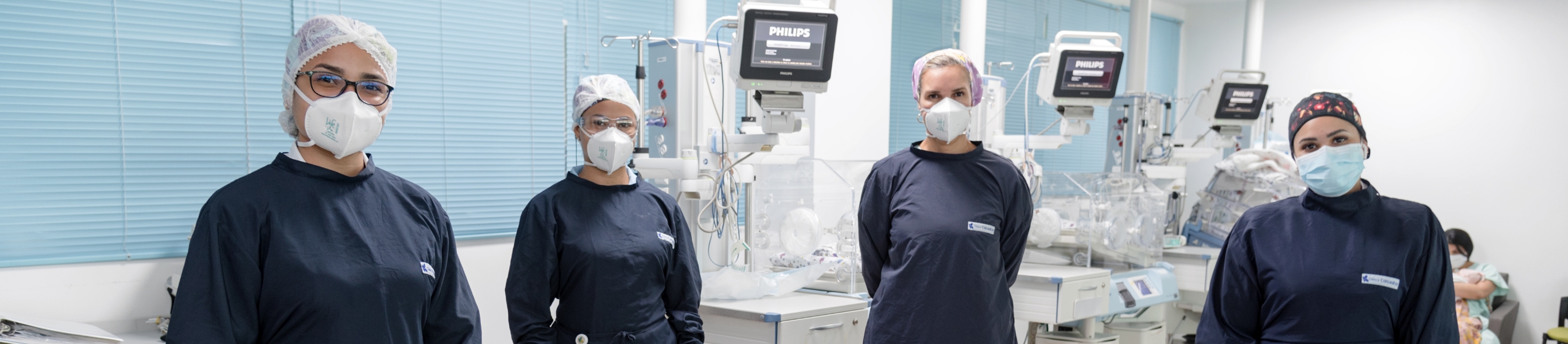 En un ambiente de UCI neonatal se ven a cuatro profesionales de la salud mirando hacía el frente, todas con trajes y gorros quirúrgicos y tapabocas. 