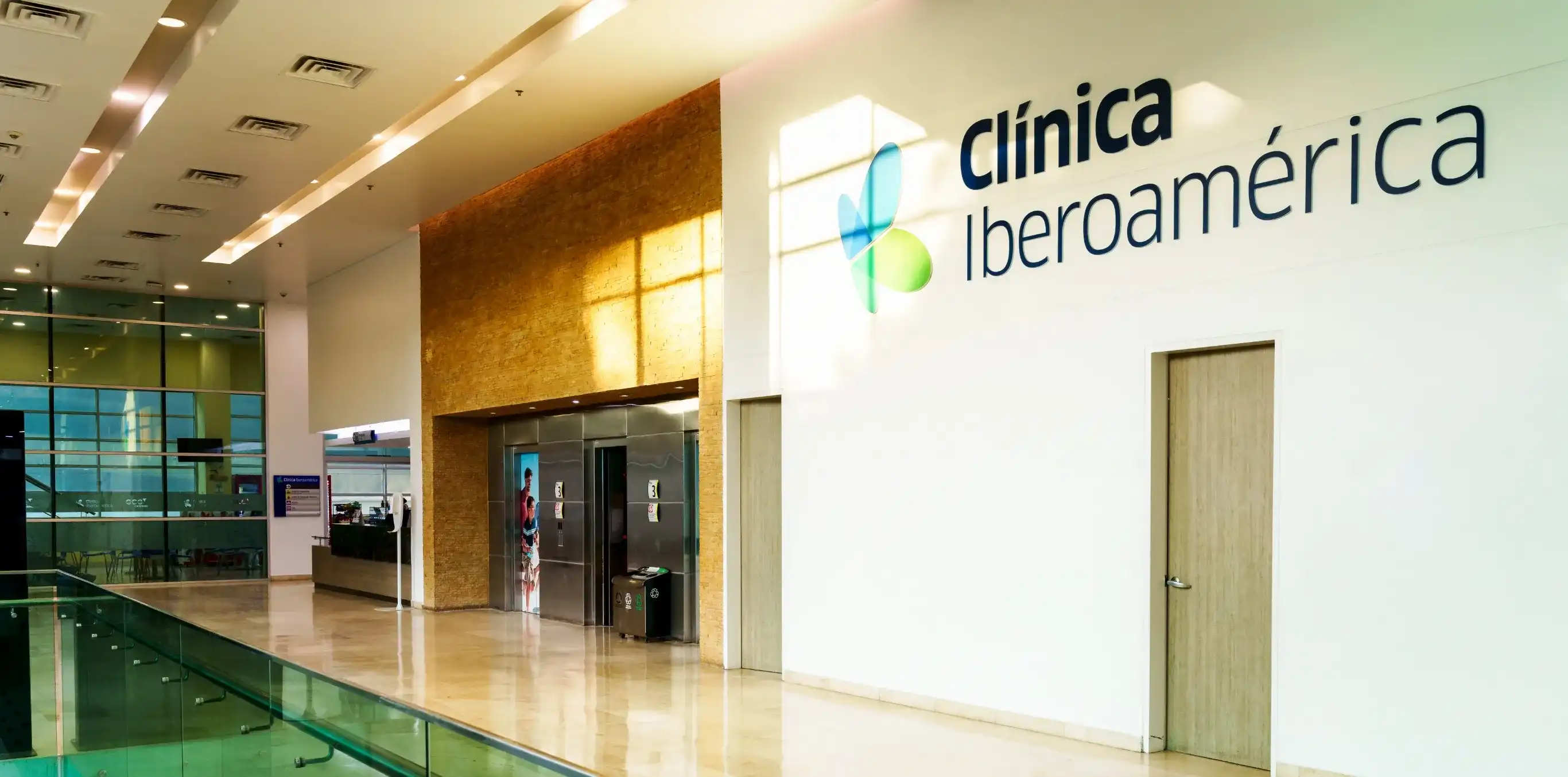En un ambiente de pasillo de hospital en la parte derecha se ve un letrero pintado en la pared que dice Clínica Iberoamérica, más adelante se ven tres ascensores.